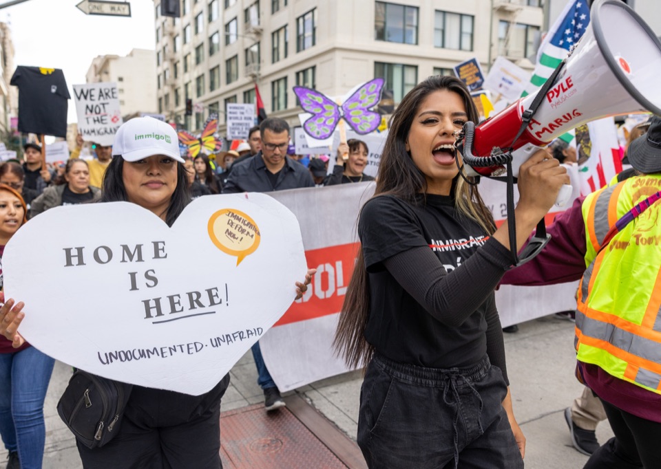 A marcher speaking through a megaphone beside a Home Is Here sign